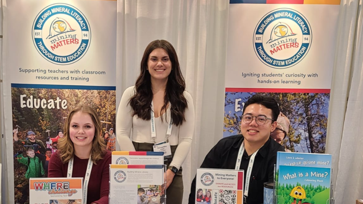 The image shows a booth setup for Mining Matters, a charitable organization focused on STEM education related to mineral literacy. Three representatives are pictured at the booth. The table features various informational materials, including brochures, pamphlets, posters and activity books for children. Behind the booth, banners display the organization's mission to support teachers with classroom resources and to ignite students' curiosity through hands-on learning.