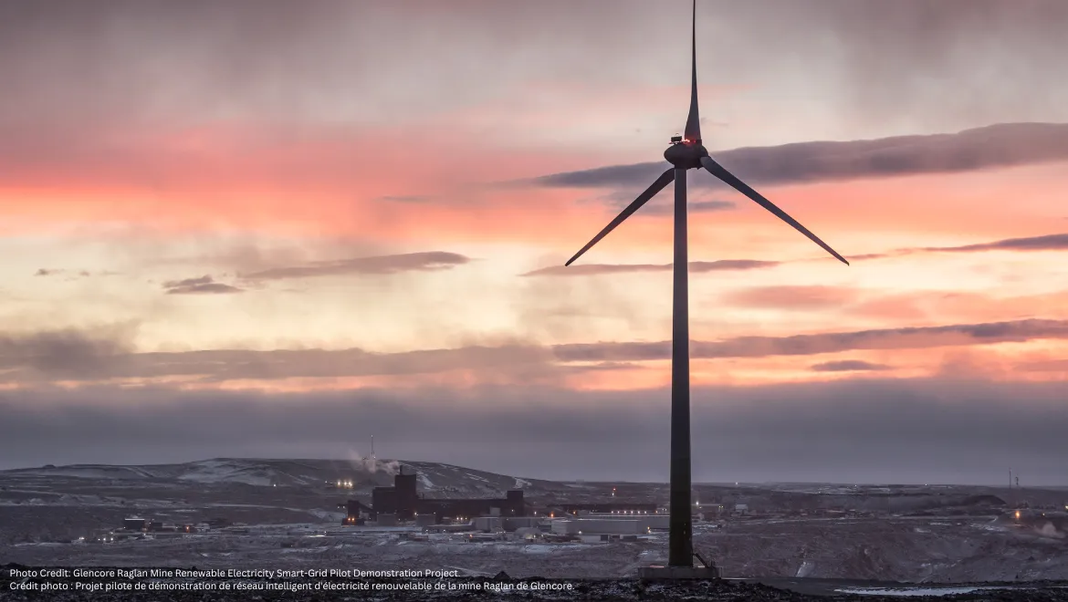 A wind turbine at Glencore's Raglan Mine in northern Quebec stands against a pink and orange sunset, with the snowy mining site in the background. Une éolienne de la mine Raglan de Glencore, dans le nord du Québec, se dresse devant un coucher de soleil rose et orange, avec le site minier enneigé en arrière-plan.