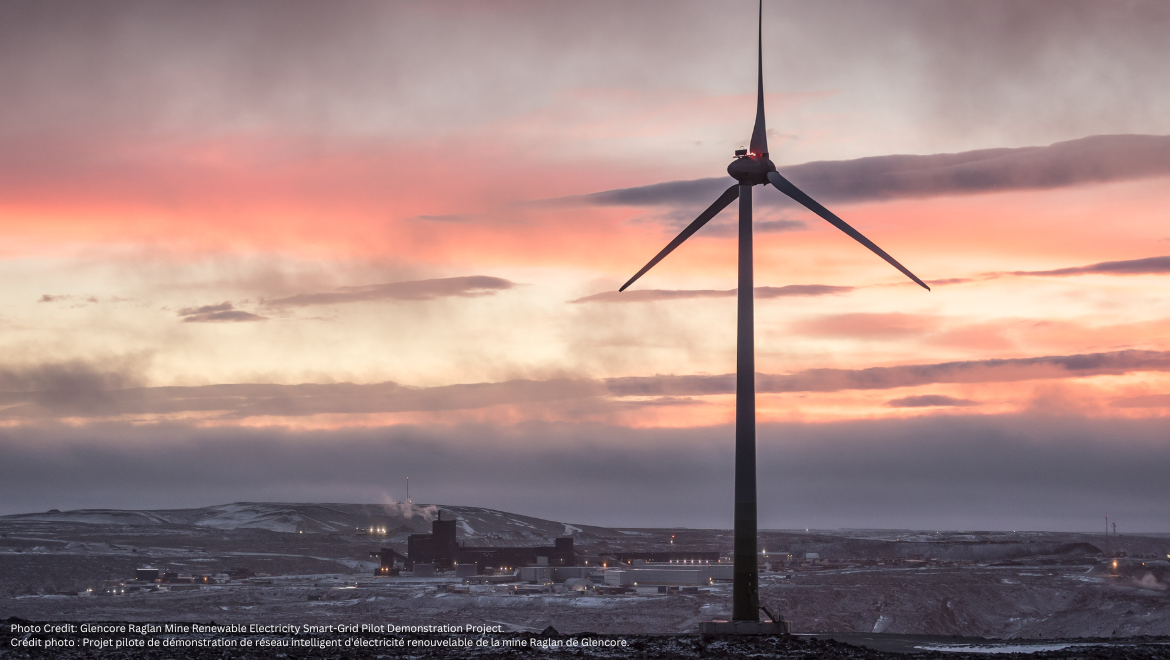 A wind turbine at Glencore's Raglan Mine in northern Quebec stands against a pink and orange sunset, with the snowy mining site in the background. Une éolienne de la mine Raglan de Glencore, dans le nord du Québec, se dresse devant un coucher de soleil rose et orange, avec le site minier enneigé en arrière-plan.
