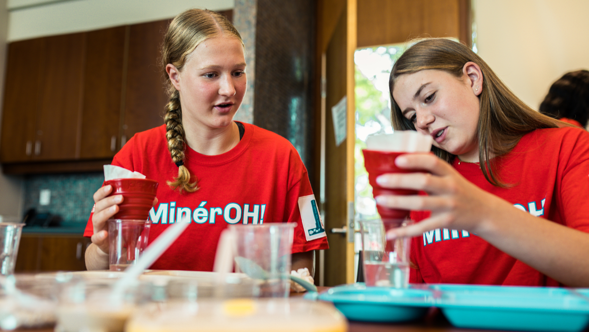 Two teenage girls wearing matching red “MinérOH!” T-shirts work together on a water filtration engineering challenge. Seated at a table covered with cups, trays, and filter materials, they concentrate on pouring water through red funnel-style filters. One girl watches attentively while the other carefully examines her filtration setup.