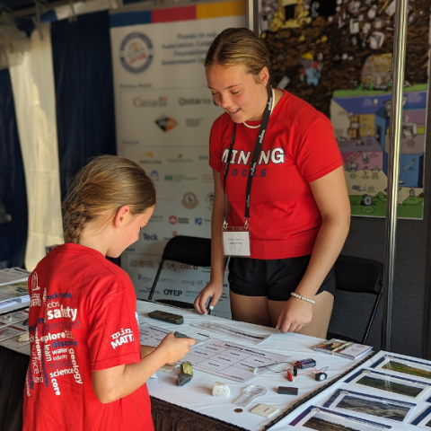 Un jeune examine attentivement un échantillon minéral à une table d'activités géologiques pratiques. La table est recouverte de divers spécimens de roches et de minéraux, d'outils d'analyse et de matériel pédagogique.