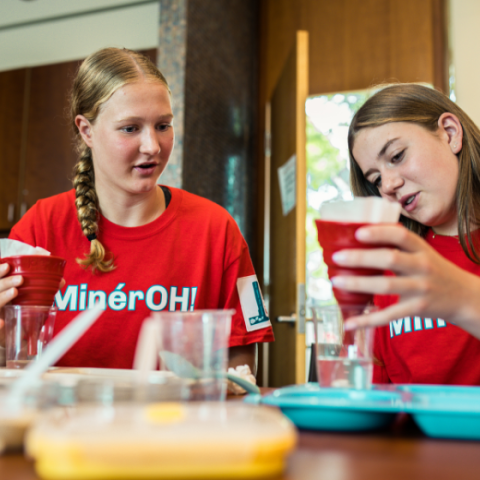Two teenage girls wearing matching red “MinérOH!” T-shirts work together on a water filtration engineering challenge. Seated at a table covered with cups, trays, and filter materials, they concentrate on pouring water through red funnel-style filters. One girl watches attentively while the other carefully examines her filtration setup.