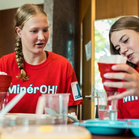 Two teenage girls wearing matching red “MinérOH!” T-shirts work together on a water filtration engineering challenge. Seated at a table covered with cups, trays, and filter materials, they concentrate on pouring water through red funnel-style filters. One girl watches attentively while the other carefully examines her filtration setup.