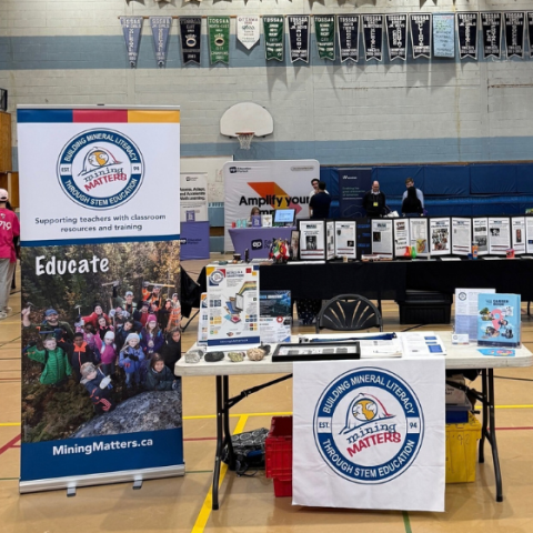 A Mining Matters educational booth in a school gymnasium, featuring a large branded banner, educational posters and materials, mineral samples, books, and resources about minerals, mining, and careers in the industry.