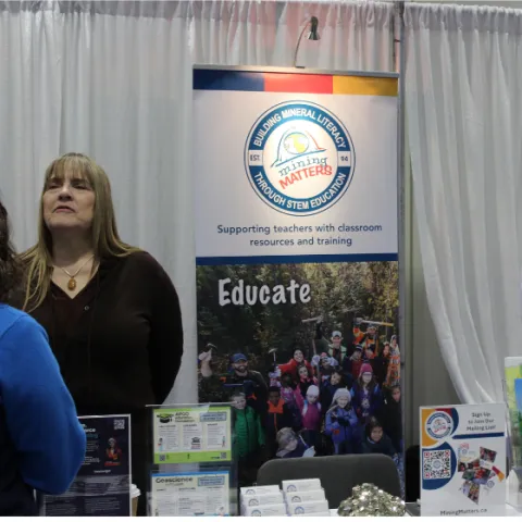 Mining Matters' booth with rock and mineral samples and educational pamphlets along with staff engaging with visitors at the event • Le kiosque de Mining Matters, avec ses échantillons de roches et de minéraux ainsi que ses brochures éducatives, et le personnel échangeant avec les visiteurs lors de l'événement.