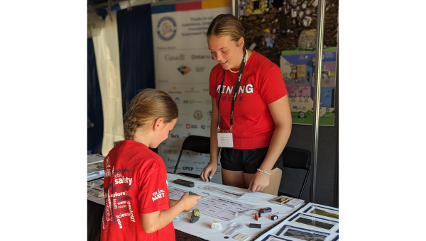 A young person is closely examining a mineral sample at a hands-on geology activity table. The table is covered with various rock and mineral specimens, testing tools, and educational materials.