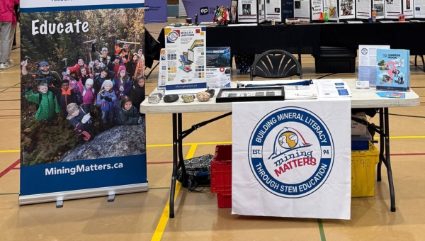 A Mining Matters educational booth in a school gymnasium, featuring a large branded banner, educational posters and materials, mineral samples, books, and resources about minerals, mining, and careers in the industry.